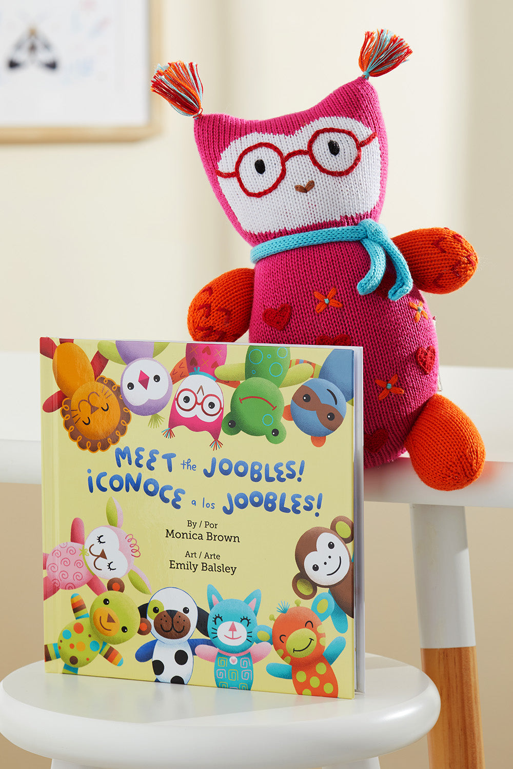 A colorful knit stuffed toy with glasses sits behind a childrens book titled Meet the Joobles! ¬°Conoce a los Joobles! placed on a white chair.