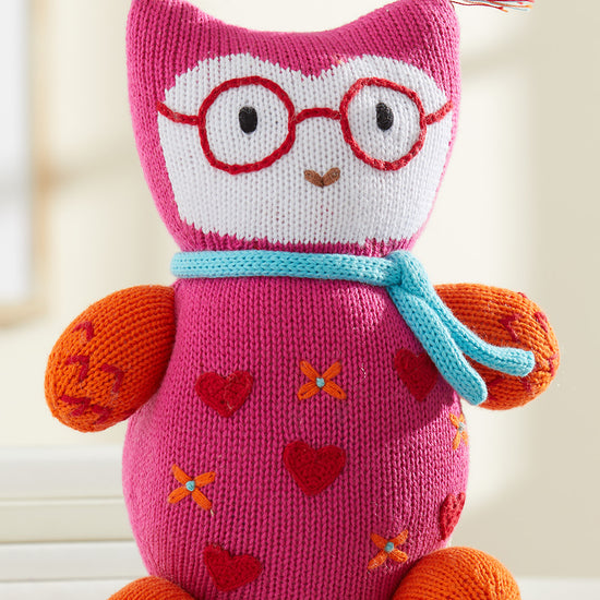 A knitted stuffed owl with red glasses, a blue scarf, orange feet and arms, pink body with heart patterns, and colorful yarn tufts on the ears.