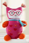 A knitted stuffed owl with red glasses, a blue scarf, orange feet and arms, pink body with heart patterns, and colorful yarn tufts on the ears.