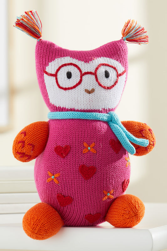 A knitted stuffed owl with red glasses, a blue scarf, orange feet and arms, pink body with heart patterns, and colorful yarn tufts on the ears.