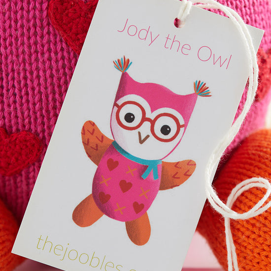 A product tag with an illustration of a colorful owl named Jody the Owl and the website thejoobles.com attached to a knitted pink and red item.