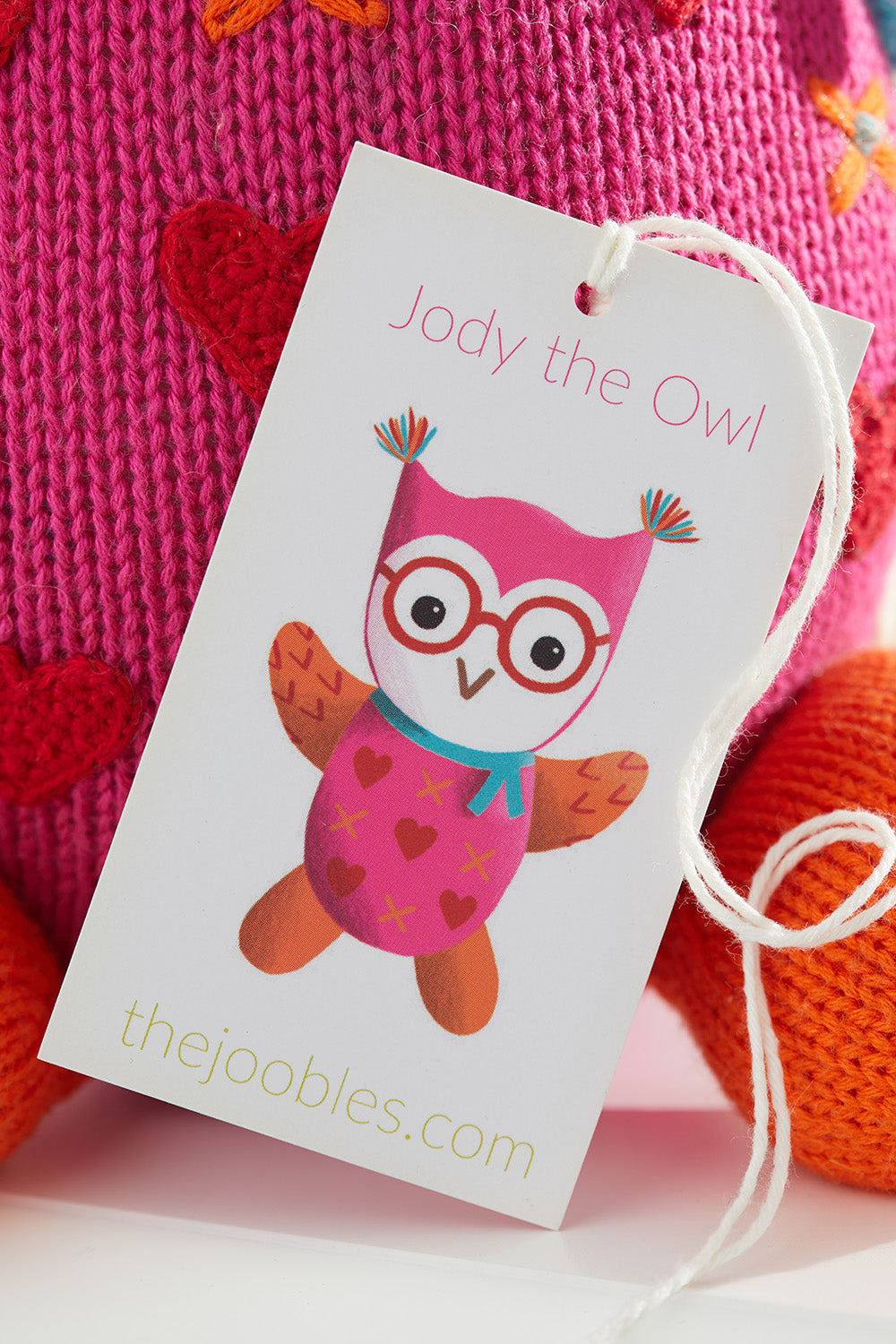 A product tag with an illustration of a colorful owl named Jody the Owl and the website thejoobles.com attached to a knitted pink and red item.