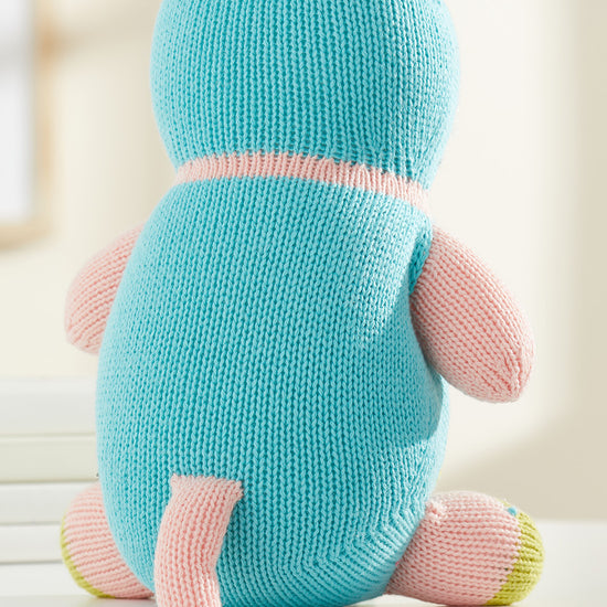 A knitted stuffed animal with blue and pink yarn sits facing away on a white surface, showing its back, ears, tail, and limbs.