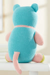 A knitted stuffed animal with blue and pink yarn sits facing away on a white surface, showing its back, ears, tail, and limbs.