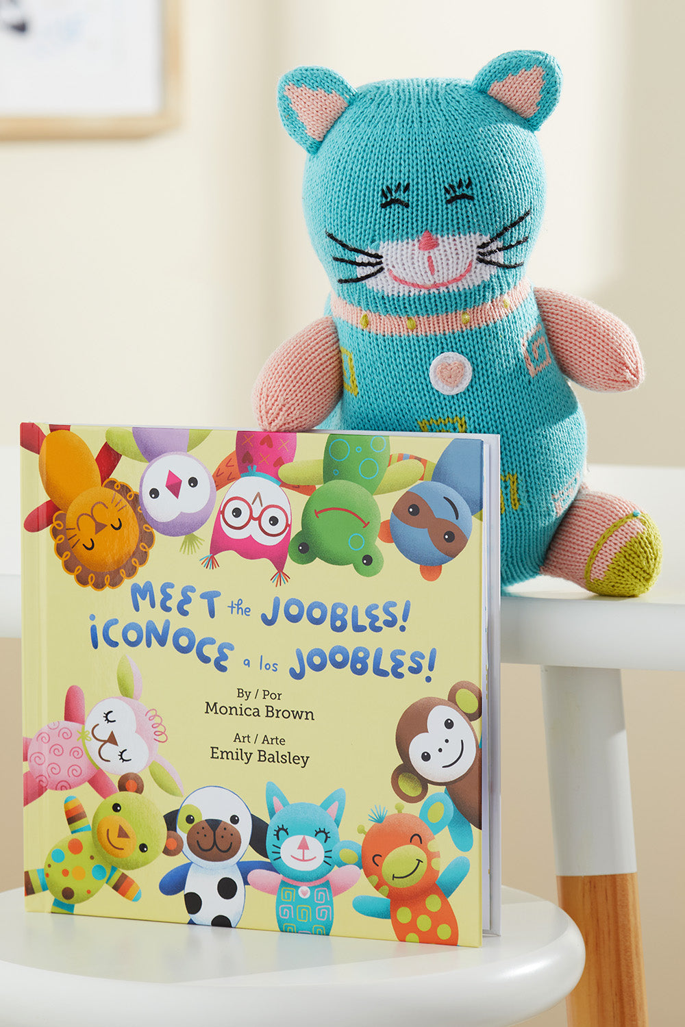 A knitted blue cat plush sits behind a bilingual children‚Äôs book titled Meet the Joobles! ¬°Conoce a los Joobles! on a white chair.
