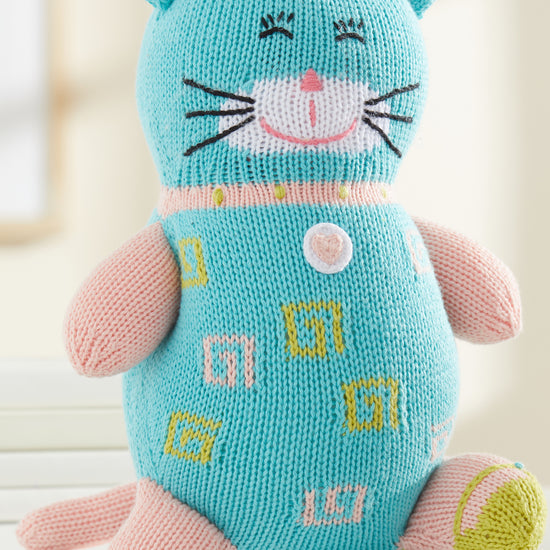 A knitted stuffed cat toy with blue and pink yarn, embroidered facial features, and patterned designs on its body, sitting upright on a white surface.
