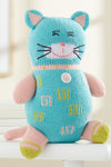 A knitted stuffed cat toy with blue and pink yarn, embroidered facial features, and patterned designs on its body, sitting upright on a white surface.