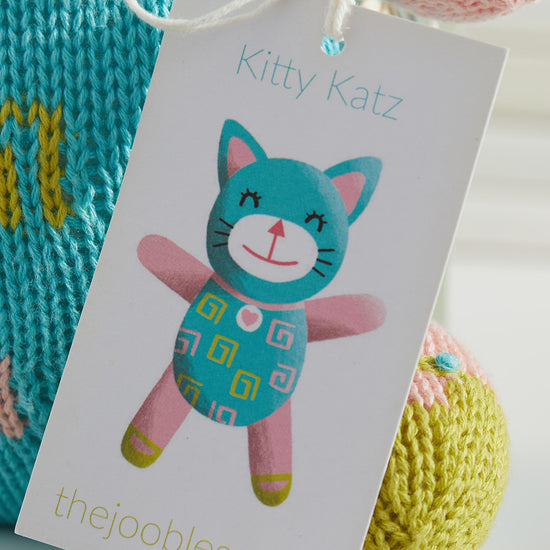 A product tag labeled Kitty Katz featuring an illustration of a teal and pink cat doll is attached to a knitted toy. The website thejoobles.com appears at the bottom of the tag.