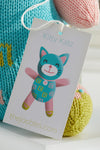 A product tag labeled Kitty Katz featuring an illustration of a teal and pink cat doll is attached to a knitted toy. The website thejoobles.com appears at the bottom of the tag.