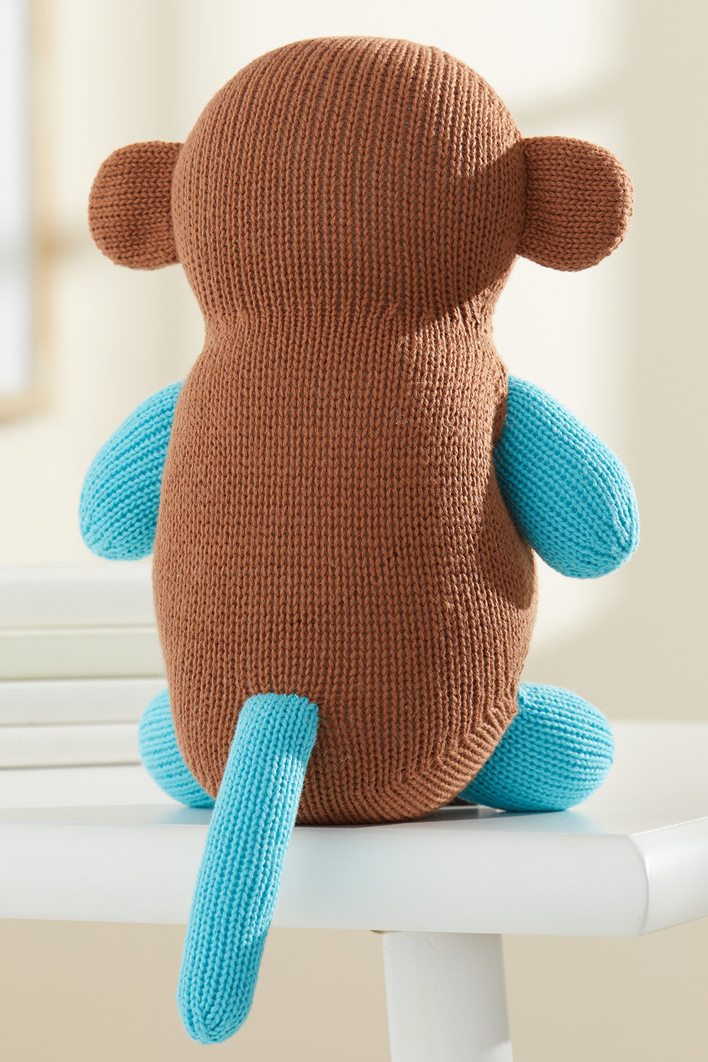 A knitted stuffed monkey toy with a brown body and blue arms, legs, and tail sits on a white surface, facing away.