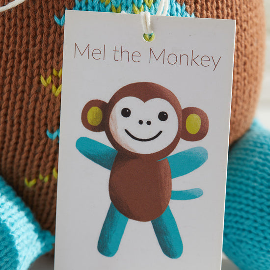 A product tag with an illustration of a smiling cartoon monkey labeled Mel the Monkey and the website thejoobles.com in front of a knitted toy.