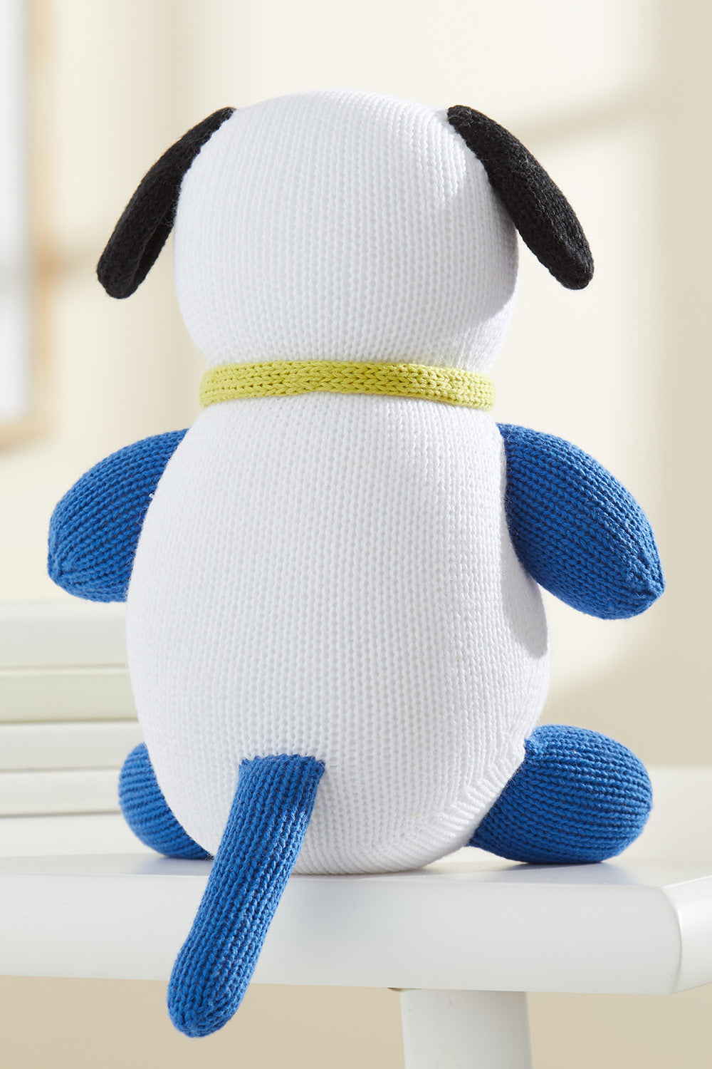 A knitted stuffed dog with a white body, blue legs and tail, black ears, and a yellow collar, sitting on a white surface facing away.