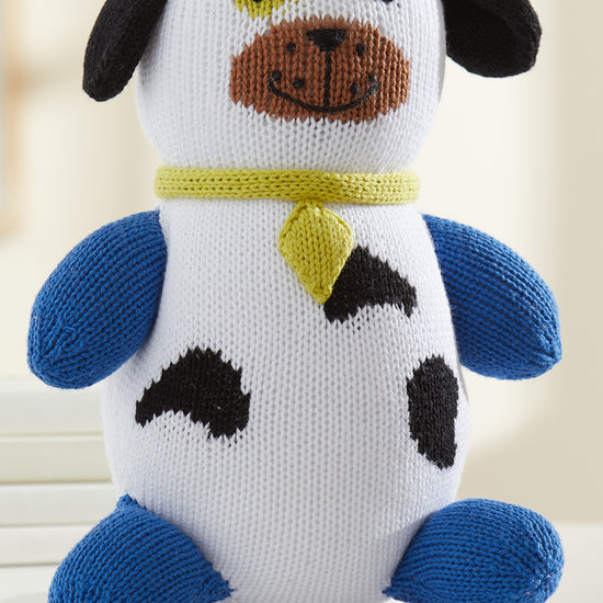 A knitted plush dog toy with blue limbs, black spots, a yellow collar, and a brown snout, sitting on a white surface.