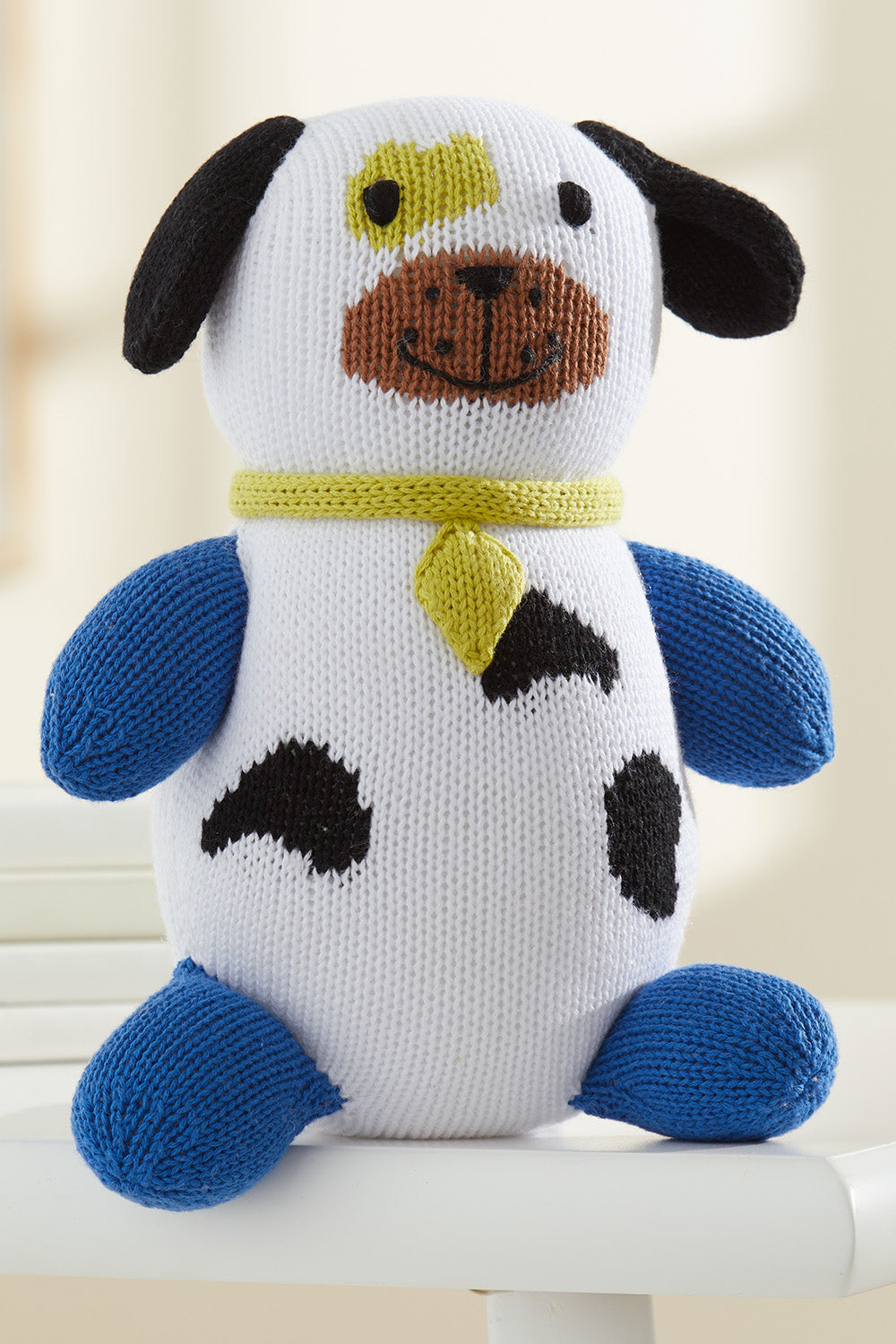 A knitted plush dog toy with blue limbs, black spots, a yellow collar, and a brown snout, sitting on a white surface.
