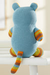 A knitted blue plush toy with striped orange, yellow, and green arms, legs, ears, and tail, shown from the back, sits on a white surface.