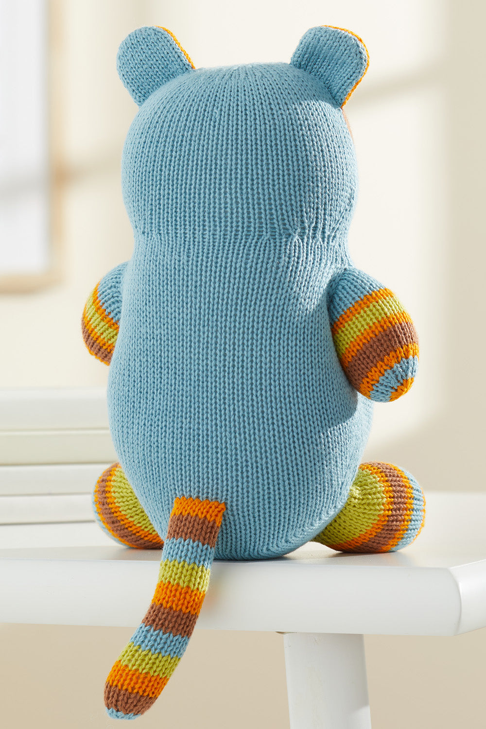 A knitted blue plush toy with striped orange, yellow, and green arms, legs, ears, and tail, shown from the back, sits on a white surface.