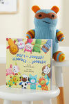 A knitted blue and orange stuffed animal sits behind a bilingual childrens book titled Meet the Joobles! ¬°Conoce a los Joobles! displayed upright on a white chair.