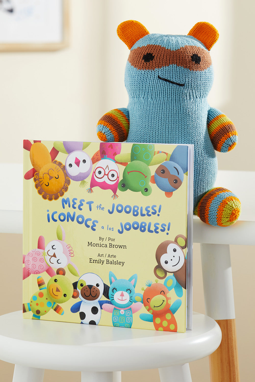 A knitted blue and orange stuffed animal sits behind a bilingual childrens book titled Meet the Joobles! ¬°Conoce a los Joobles! displayed upright on a white chair.