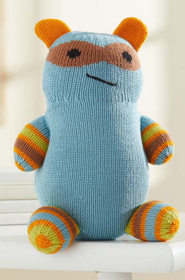 A knitted stuffed toy with blue body, orange and yellow striped limbs, orange ears, and a brown mask-like pattern on its face sits on a white chair.