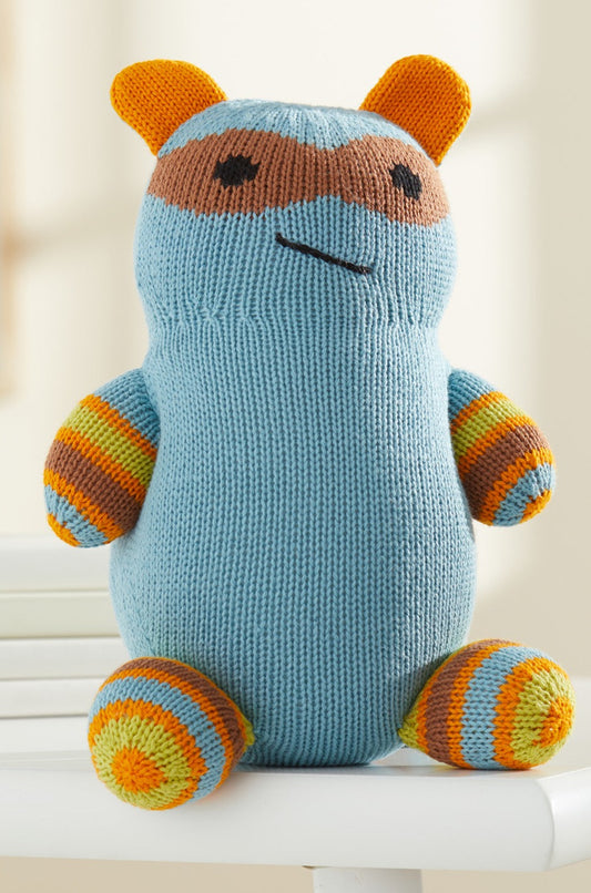 A knitted stuffed toy with blue body, orange and yellow striped limbs, orange ears, and a brown mask-like pattern on its face sits on a white chair.
