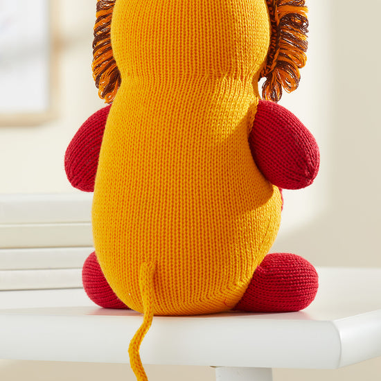 A knitted lion plush with a yellow body, red limbs, and a brown and yellow mane sits on a white surface facing away.