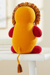 A knitted lion plush with a yellow body, red limbs, and a brown and yellow mane sits on a white surface facing away.