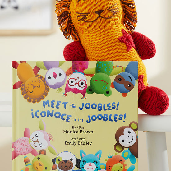 A knit lion plush toy sits behind a childrens book titled Meet the Joobles! ¬°Conoce a los Joobles! featuring colorful illustrated animal characters on the cover.