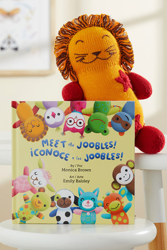 A knit lion plush toy sits behind a childrens book titled Meet the Joobles! ¬°Conoce a los Joobles! featuring colorful illustrated animal characters on the cover.