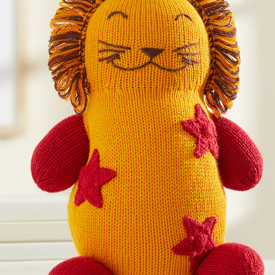 A knitted stuffed lion toy with a yellow body, red arms and legs, red star patterns, and a mane made of orange and brown yarn.
