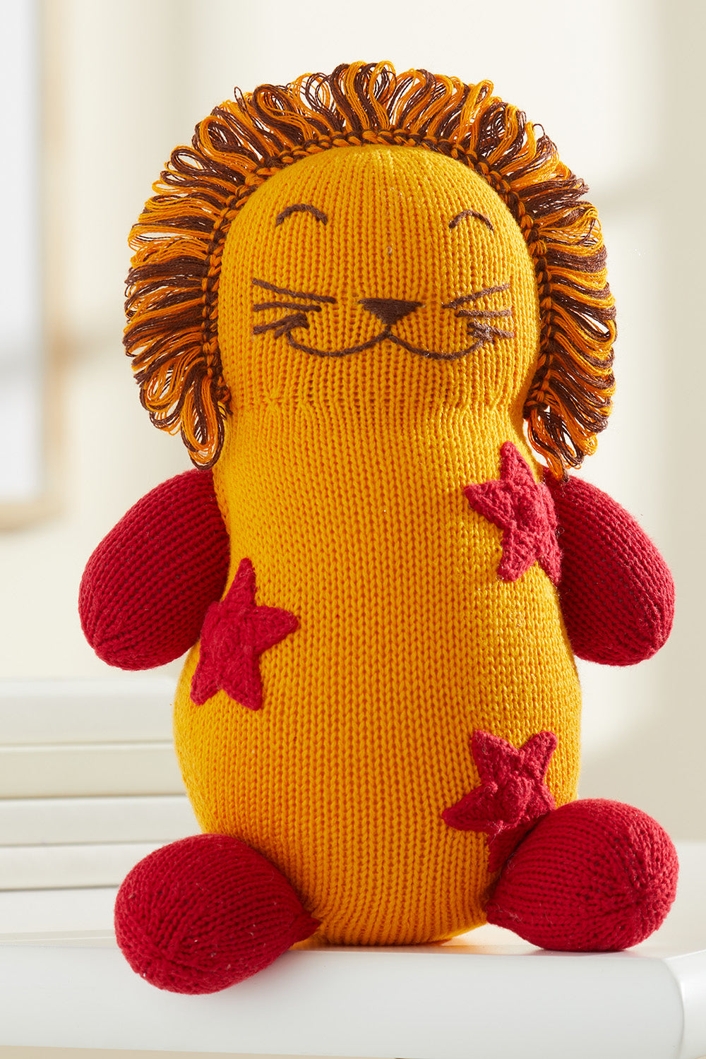 A knitted stuffed lion toy with a yellow body, red arms and legs, red star patterns, and a mane made of orange and brown yarn.