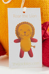 A product tag with an illustration of a smiling lion named Roar the Lion and thejoobles.com printed at the bottom, attached to a yellow and red knitted item.