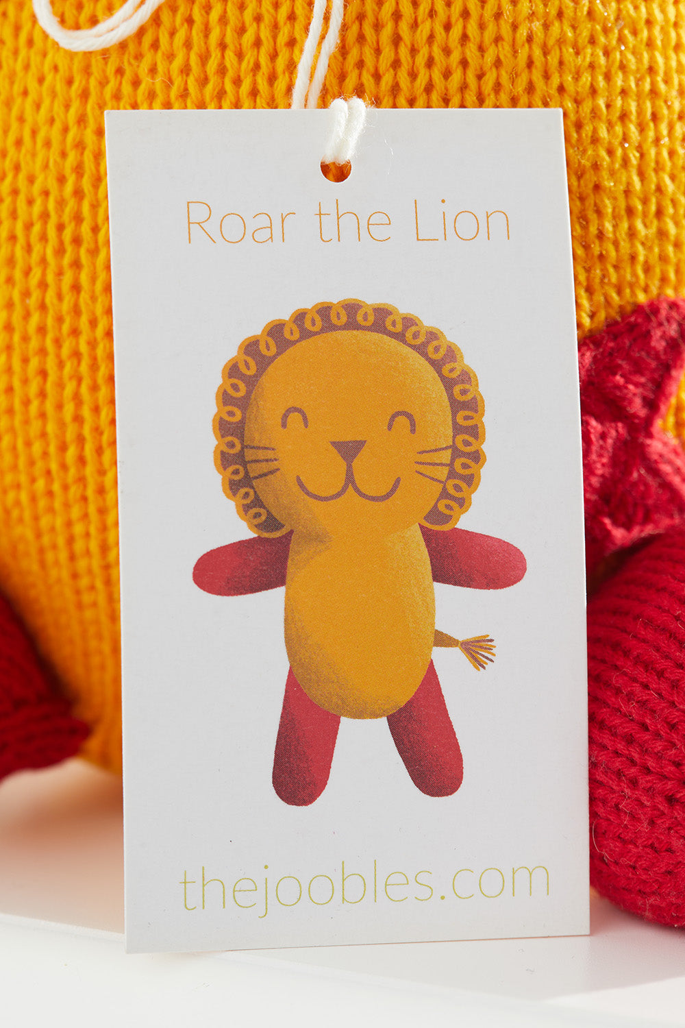A product tag with an illustration of a smiling lion named Roar the Lion and thejoobles.com printed at the bottom, attached to a yellow and red knitted item.