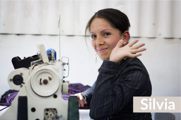 A woman sits at a sewing machine, smiling and waving at the camera. The name Silvia is displayed in white text at the bottom right.