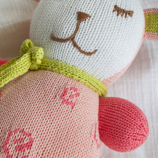A close-up of a knitted stuffed animal with a white and pink body, embroidered facial features, green knitted bow, and pink accents on the ears and arms.