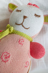 A close-up of a knitted stuffed animal with a white and pink body, embroidered facial features, green knitted bow, and pink accents on the ears and arms.