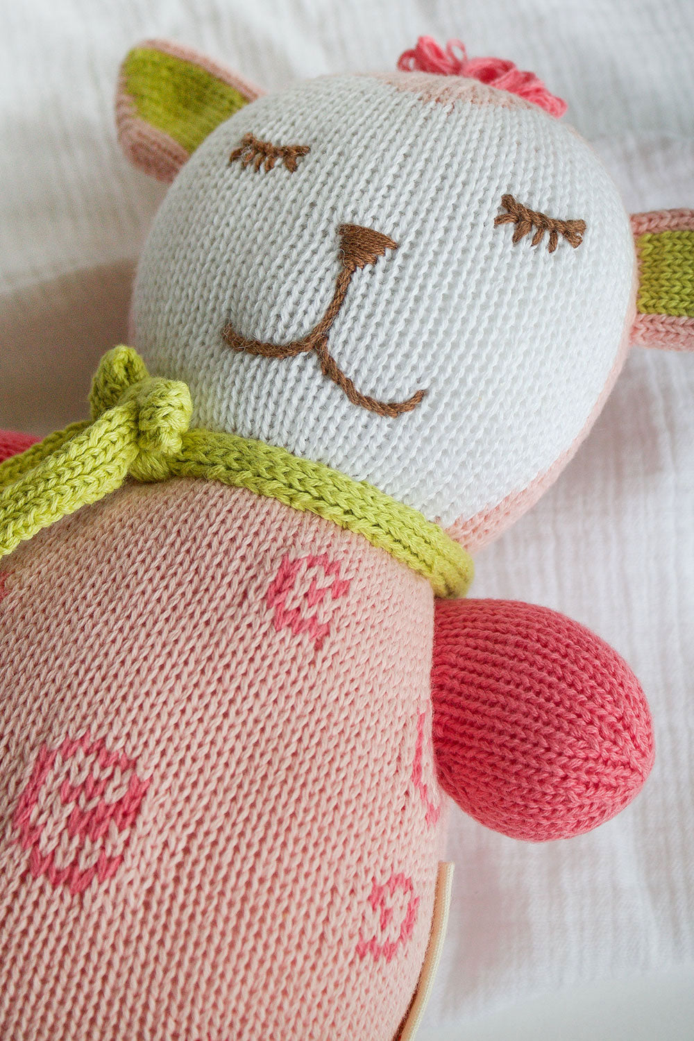A close-up of a knitted stuffed animal with a white and pink body, embroidered facial features, green knitted bow, and pink accents on the ears and arms.