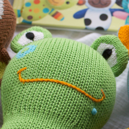 A close-up of a green knitted toy frog with embroidered eyes and a frowning orange mouth, with a childrens book in the background.