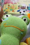 A close-up of a green knitted toy frog with embroidered eyes and a frowning orange mouth, with a childrens book in the background.