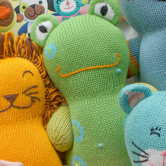 A group of colorful, knitted animal toys, including a green frog, a yellow lion, and a blue mouse, in front of a childrens book cover.