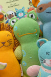 A group of colorful, knitted animal toys, including a green frog, a yellow lion, and a blue mouse, in front of a childrens book cover.