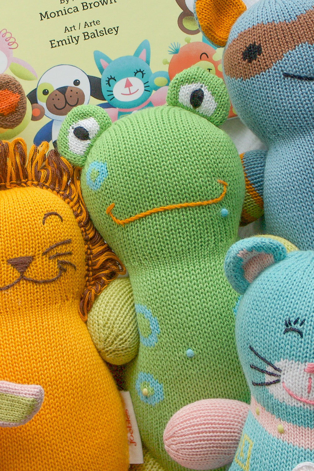 A group of colorful, knitted animal toys, including a green frog, a yellow lion, and a blue mouse, in front of a childrens book cover.