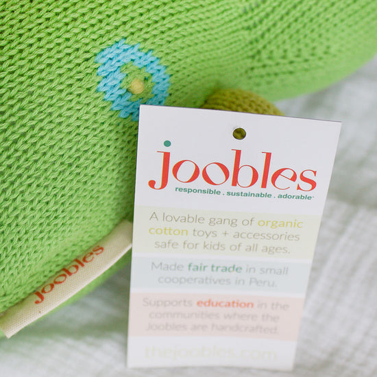 A close-up of a green knitted toy with a joobles tag, featuring a product card describing the toy as organic, fair trade, and handcrafted in Peru.