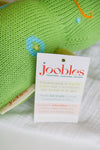 A close-up of a green knitted toy with a joobles tag, featuring a product card describing the toy as organic, fair trade, and handcrafted in Peru.