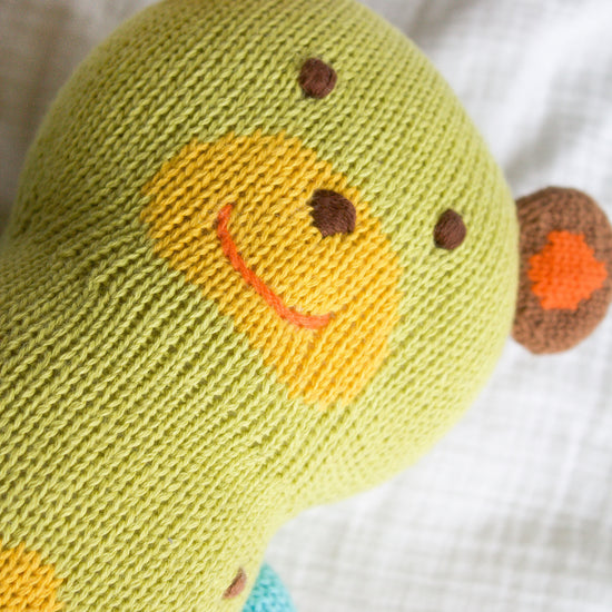 A knitted green teddy bear with a yellow snout, red smile, and brown and orange ears lies on a white fabric surface.