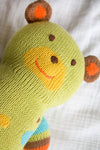 A knitted green teddy bear with a yellow snout, red smile, and brown and orange ears lies on a white fabric surface.