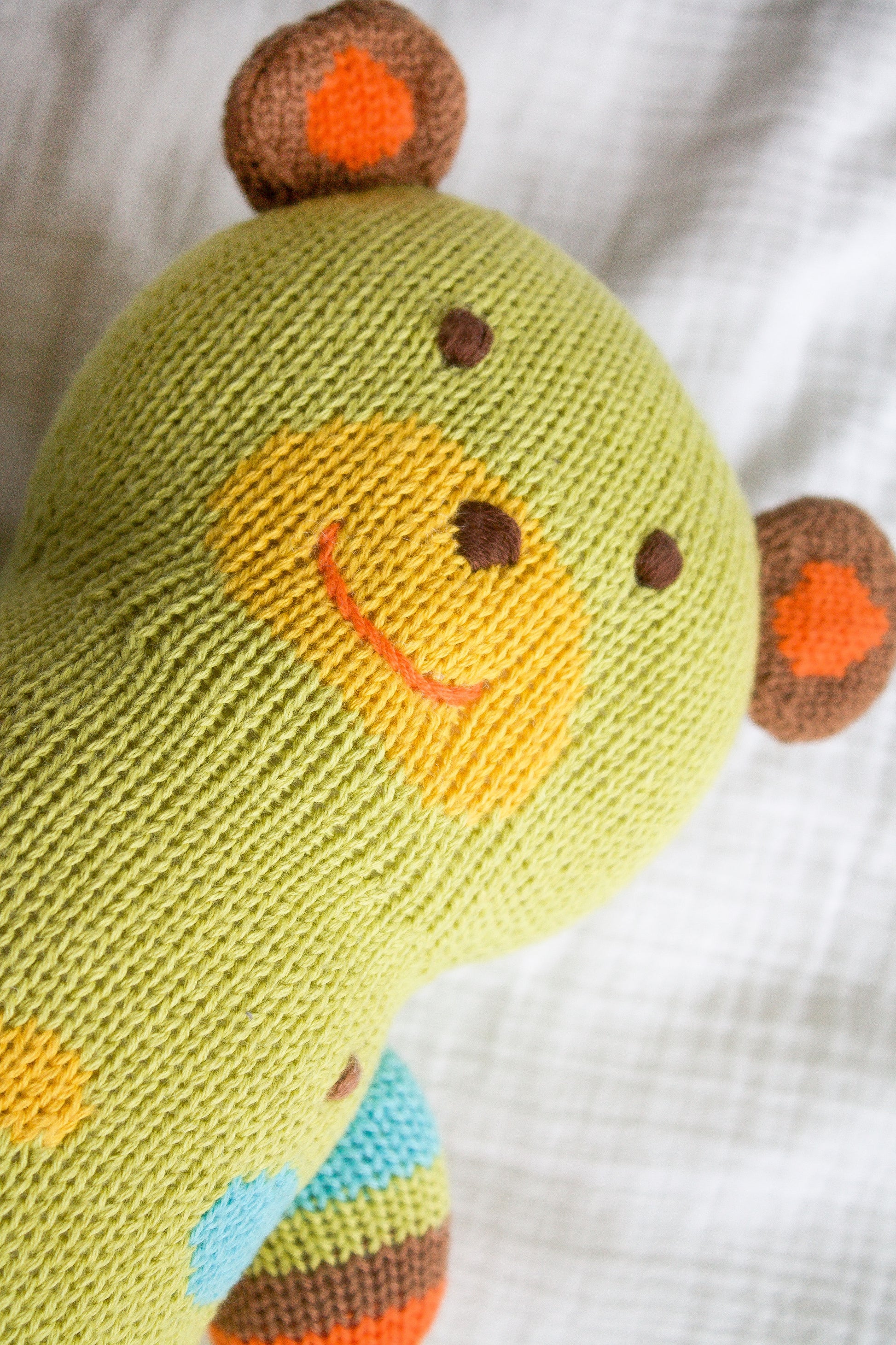 A knitted green teddy bear with a yellow snout, red smile, and brown and orange ears lies on a white fabric surface.