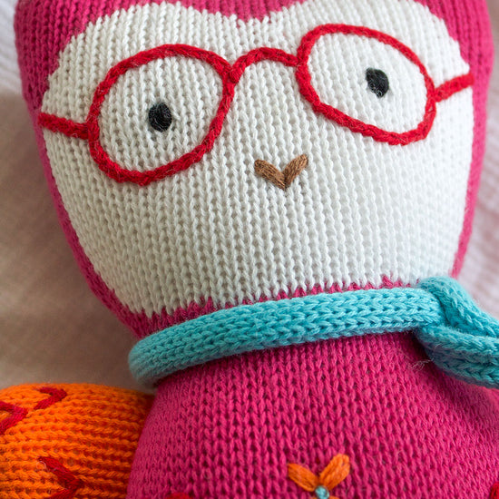 A knitted stuffed animal with red glasses, a blue scarf, and orange and red accents lies on a white fabric background.