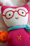 A knitted stuffed animal with red glasses, a blue scarf, and orange and red accents lies on a white fabric background.