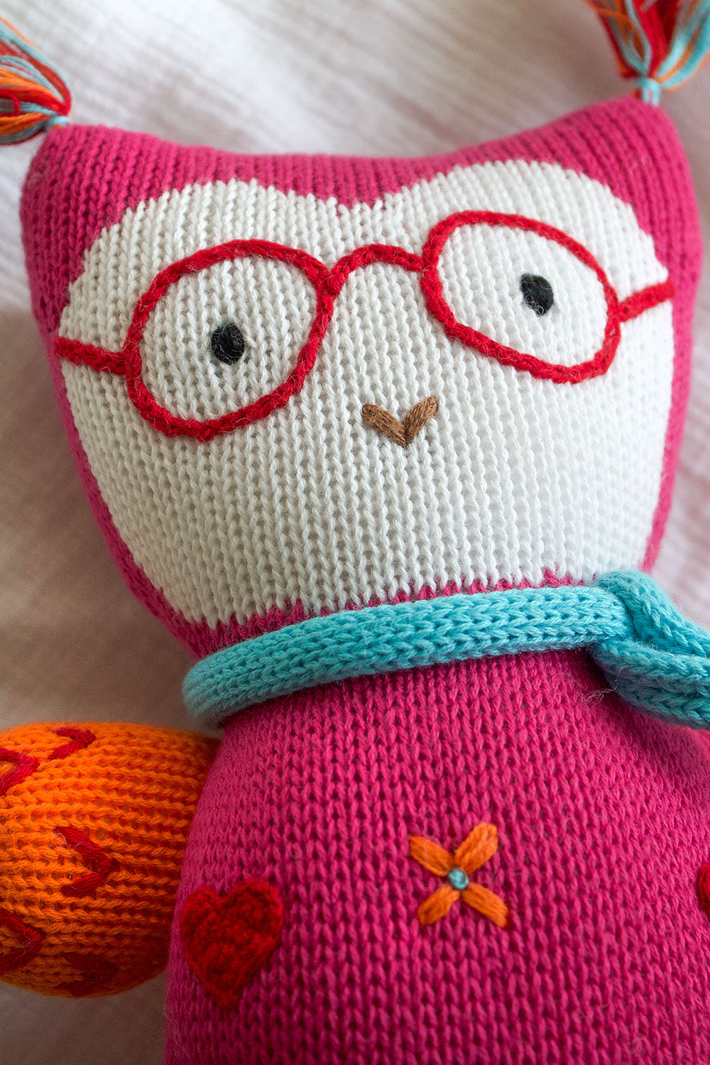 A knitted stuffed animal with red glasses, a blue scarf, and orange and red accents lies on a white fabric background.
