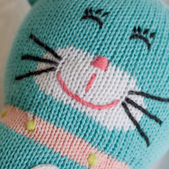 A close-up of a knitted toy cat with turquoise, white, and pink yarn, featuring stitched facial features and a heart detail on its chest.